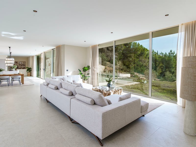 Grand salon moderne avec un canapé gris et des baies vitrées donnant sur une forêt verdoyante.