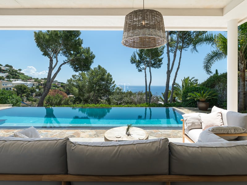 Piscine à débordement et salon extérieur sous une terrasse ombragée surplombant la mer et des pins luxuriants.
