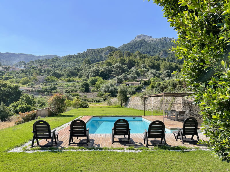 Piscine rectangulaire avec terrasse en bois et transats face aux montagnes verdoyantes sous un ciel bleu azur.