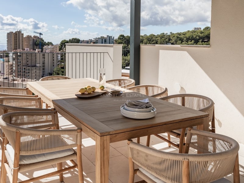 Terrasse moderne avec table en bois, chaises tressées et vue panoramique sur la mer et la ville.