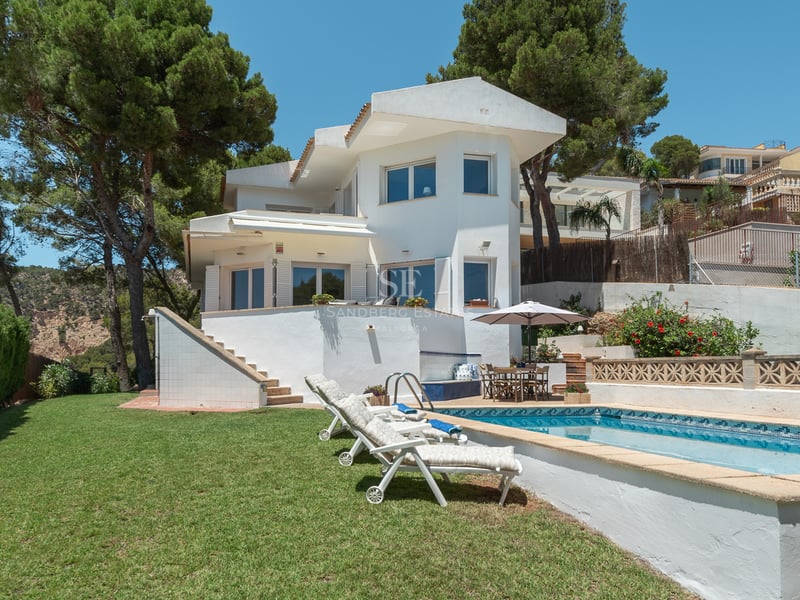 Villa blanche de deux étages avec piscine, transats et pelouse entourée de pins.