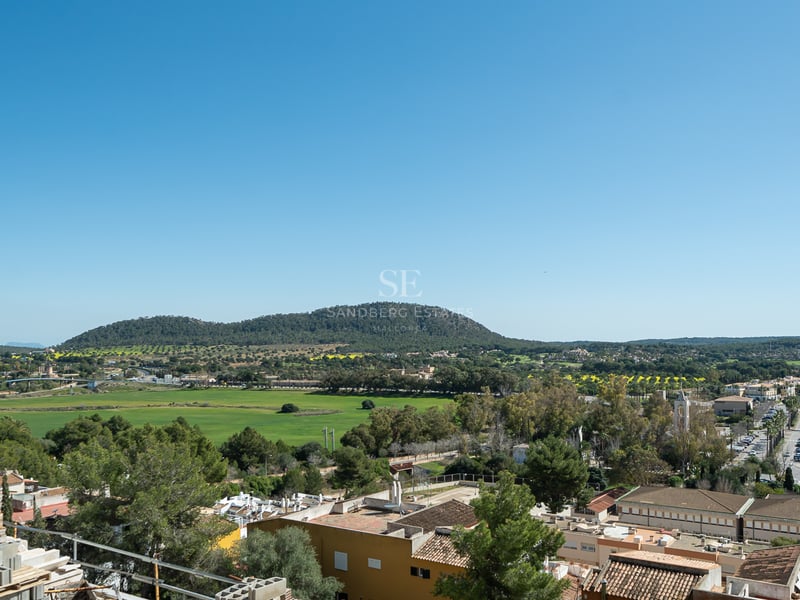 Vue en plongée d'une vallée verdoyante, de montagnes lointaines et d'un village méditerranéen sous un ciel bleu clair.