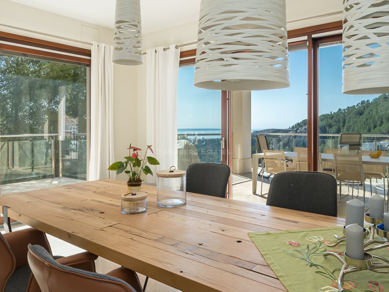 Salle à manger moderne avec table en bois rustique, chaises en cuir et baies vitrées donnant sur la côte.