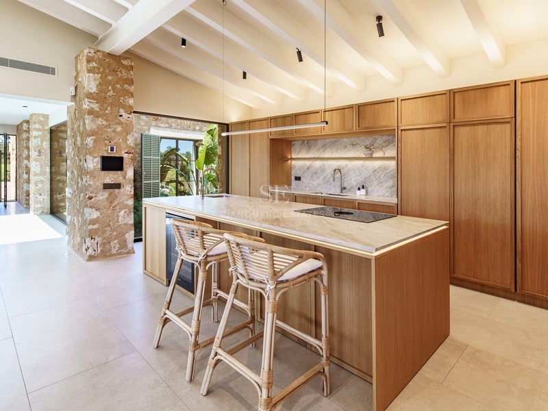 Cocina moderna con isla de madera, encimera de mármol, taburetes de mimbre y techos con vigas blancas.