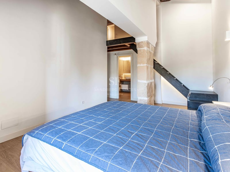 Chambre moderne avec lit bleu, pilier en pierre apparente et escalier menant à une mezzanine.