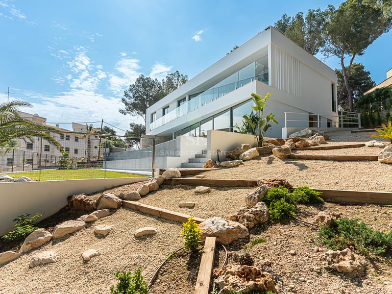 Vue latérale d'une villa blanche moderne de deux étages avec de grandes baies vitrées et un jardin de rocailles.
