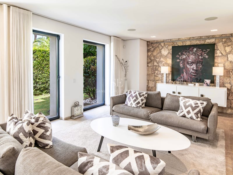 Salon lumineux avec canapés gris, mur d'accent en pierre et baies vitrées donnant sur le jardin.