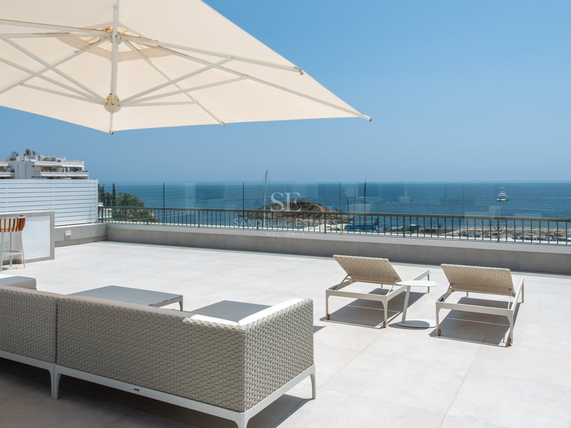 Grande terrasse ensoleillée avec mobilier de salon moderne, garde-corps en verre et vue sur la Méditerranée.
