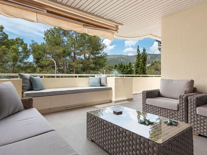 Terrasse couverte avec mobilier en osier gris, banc intégré et vue sur les pins et les montagnes.