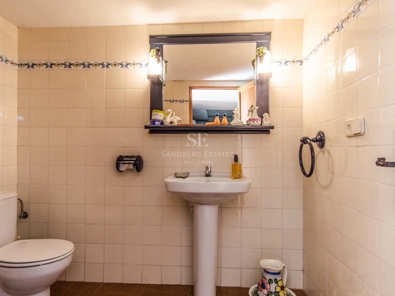 Salle de bain carrelée en crème avec lavabo sur colonne, toilettes et miroir à cadre sombre avec éclairage intégré.