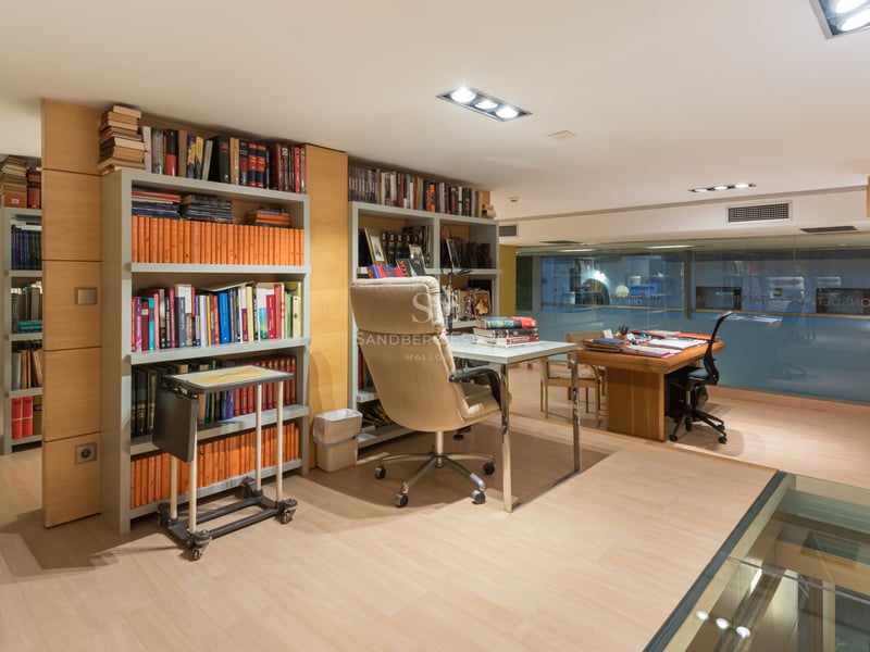 Modern home office with light wood floors, built-in bookshelves, glass desk, and ergonomic leather chair.