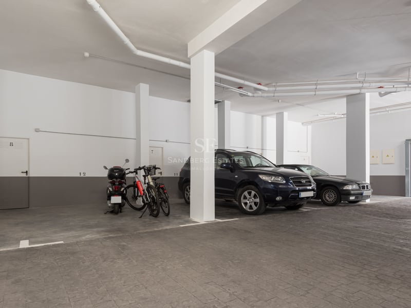 Parking souterrain avec piliers blancs, sol en pierre grise et voitures garées sous un éclairage artificiel.
