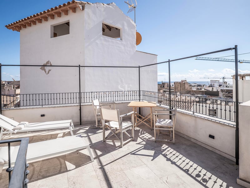 Terrasse ensoleillée avec transats blancs, table en bois et vue lointaine sur la mer au-dessus des toits.