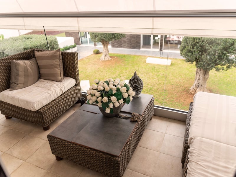 Terrasse couverte avec salon en osier, garde-corps en verre et vue sur un jardin avec oliviers.