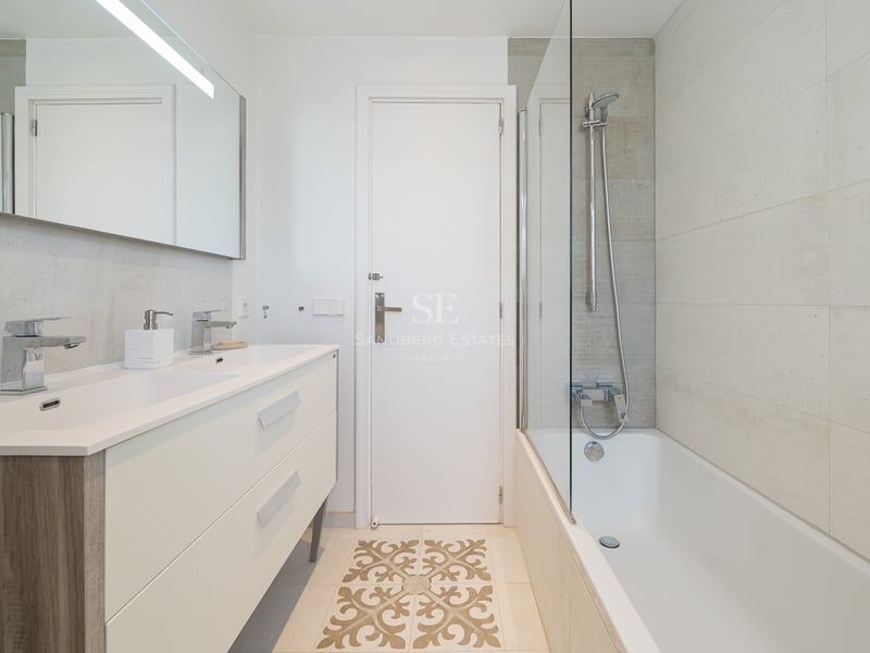 Salle de bain moderne avec double vanité crème, baignoire avec paroi vitrée et carrelage décoratif au sol.