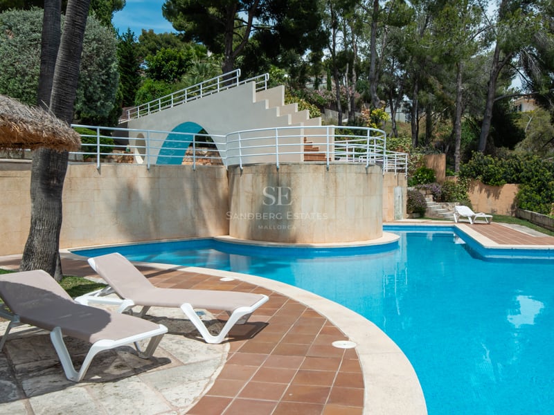 Grande piscine carrelée bleue avec chaises longues, parasol en paille et structure blanche dans un jardin arboré.