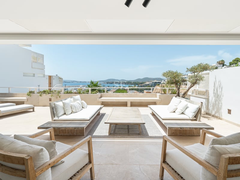 Grande terrasse blanche avec canapés et fauteuils en bois surplombant la mer bleue et la côte par une journée ensoleillée.