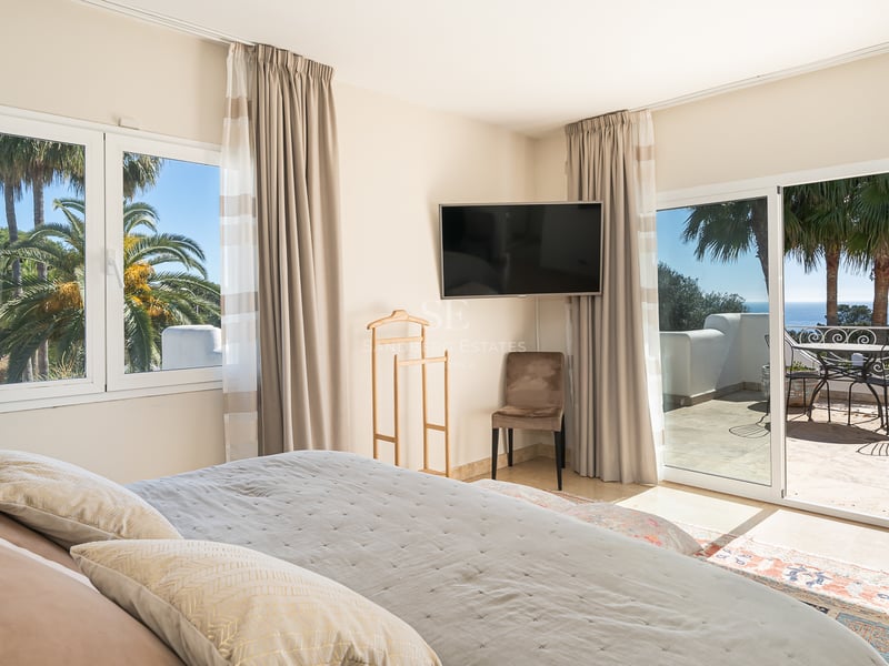 Chambre principale moderne avec grand lit, TV murale et baies vitrées ouvrant sur une terrasse ensoleillée vue mer.