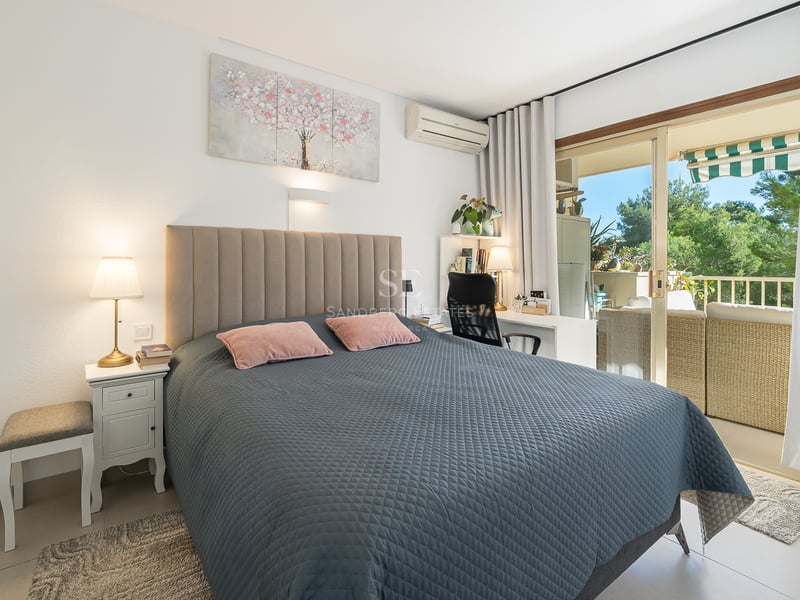 Chambre lumineuse avec lit matelassé gris, bureau et baies vitrées coulissantes ouvrant sur un balcon ensoleillé.