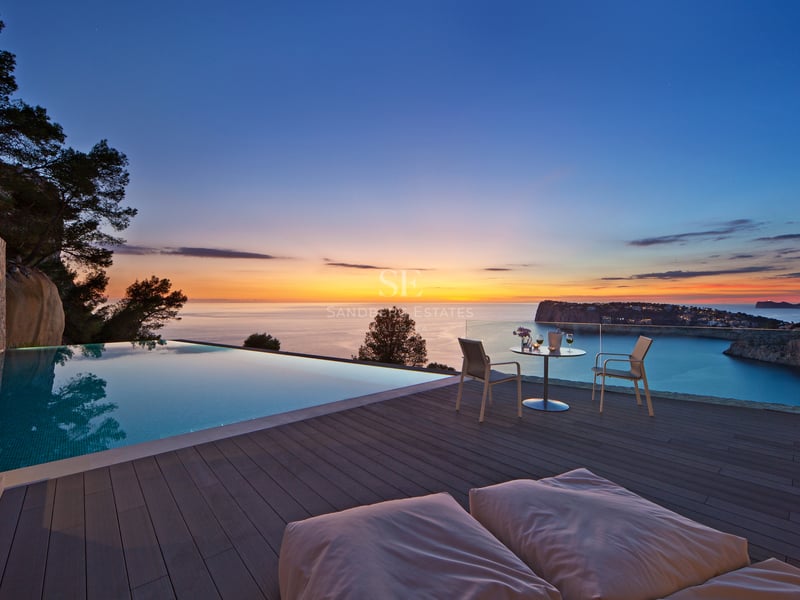 Piscine à débordement sur terrasse en bois avec vue sur la mer au coucher du soleil et mobilier de jardin.