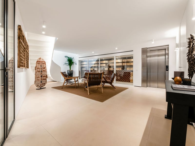 Hall minimaliste avec cave à vin vitrée, ascenseur en acier inoxydable, escalier en colimaçon blanc et fauteuils en cuir.