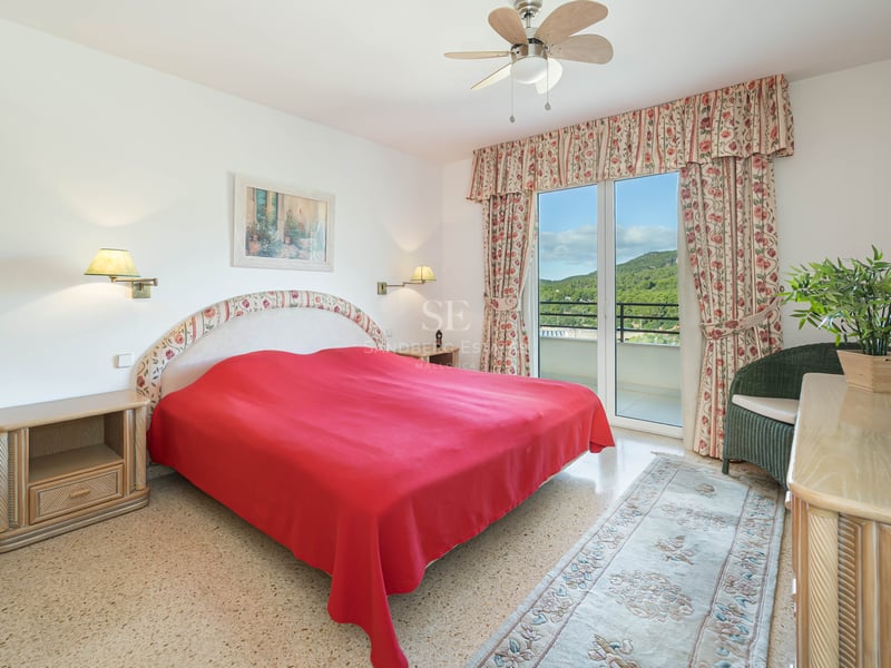 Chambre lumineuse avec couvre-lit rouge, rideaux floraux et balcon offrant une vue sur la montagne.