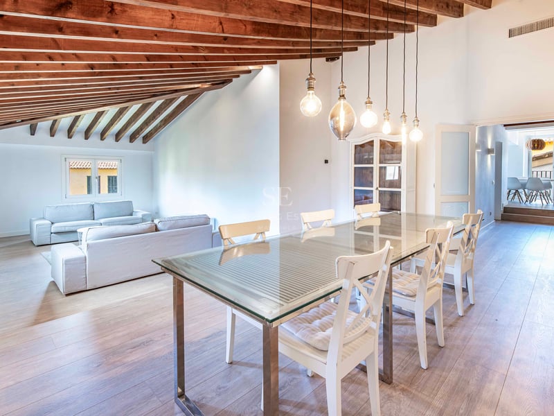 Salle à manger ouverte avec table en verre, poutres en bois au plafond et coin salon en arrière-plan.