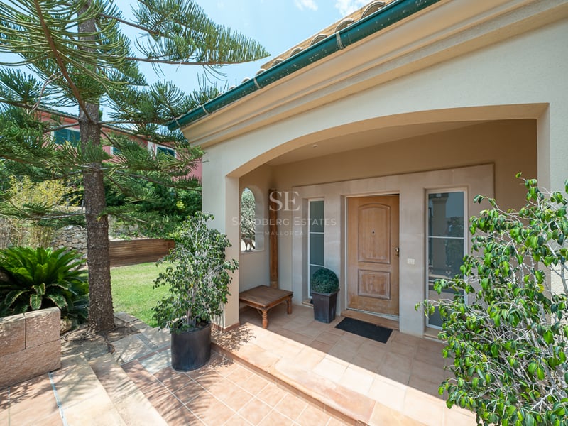 Entrée de villa voûtée avec porte en bois, sol en terre cuite et végétation luxuriante sous un ciel dégagé.