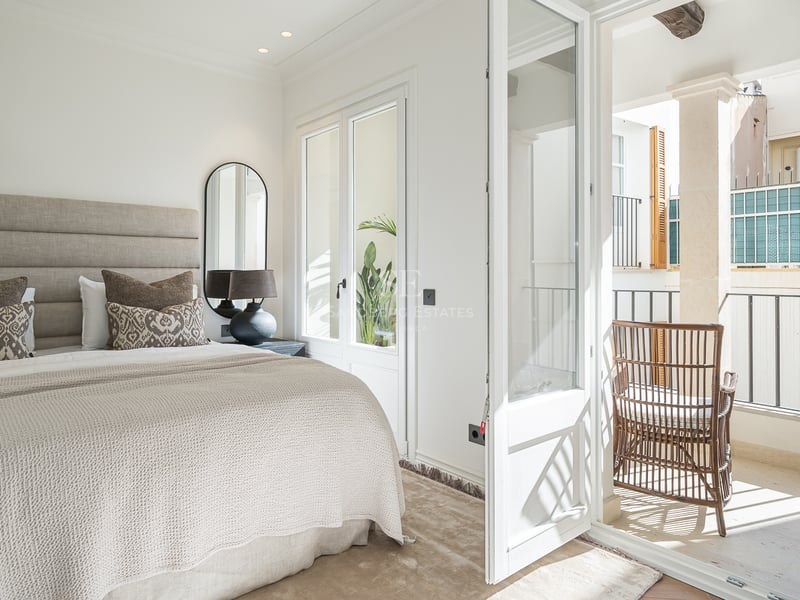 Bright bedroom with neutral linens, upholstered headboard, and open glass doors leading to a stone-tiled balcony.
