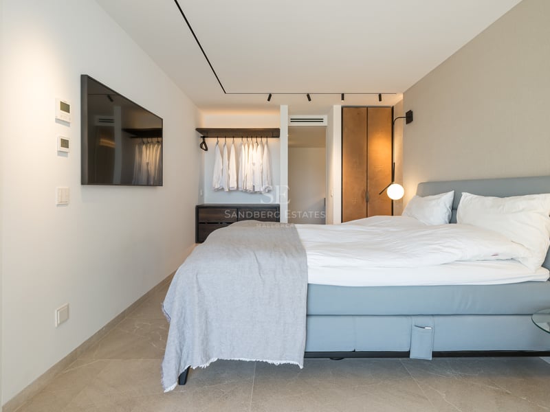 Chambre moderne avec lit bleu, draps blancs, TV murale et dressing ouvert avec éclairage sur rail.
