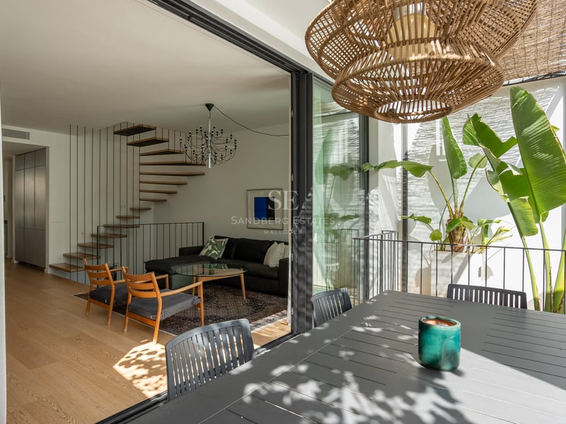 Salon moderne avec escalier en bois vu depuis une terrasse avec plantes tropicales à travers des baies vitrées.