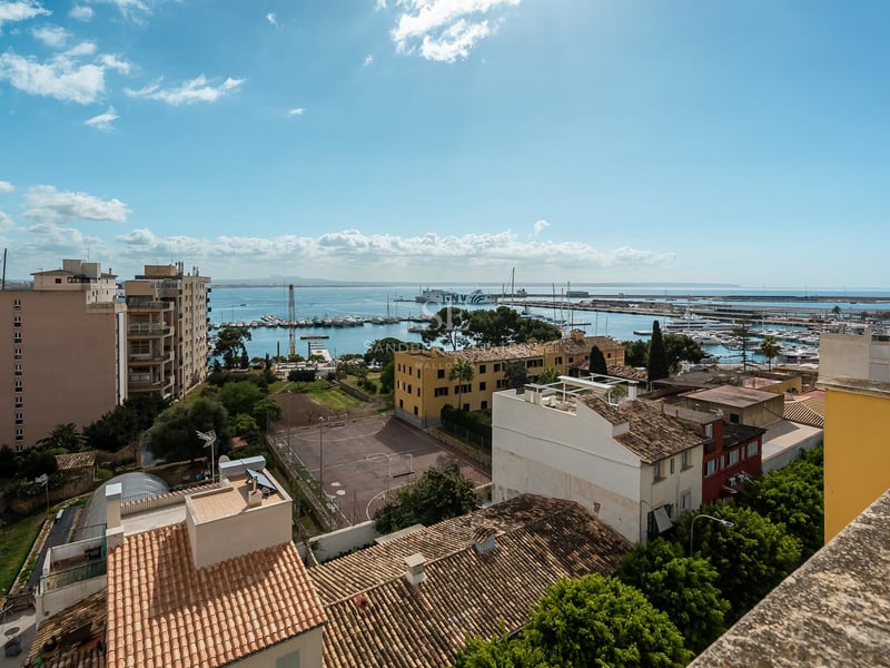 Vue surélevée d'un port méditerranéen avec des yachts, des toits en terre cuite et la mer bleue sous un ciel clair.