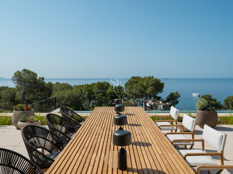 Une table à manger en bois sur une terrasse ensoleillée surplombant la mer Méditerranée et des pins.