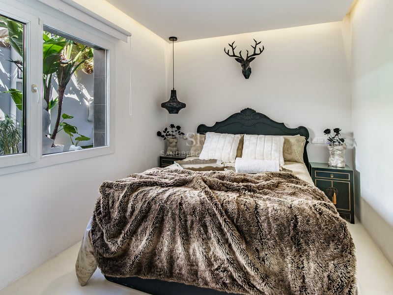 Chambre avec jeté de lit en fourrure, tête de lit sombre, tête de cerf décorative et fenêtre sur verdure.
