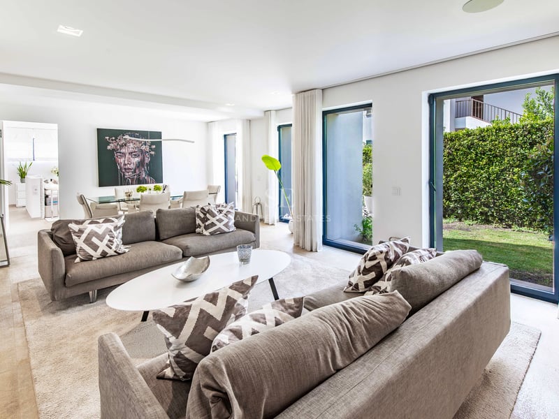 Salon lumineux avec deux canapés gris, table basse blanche et grandes baies vitrées ouvrant sur un jardin privé.