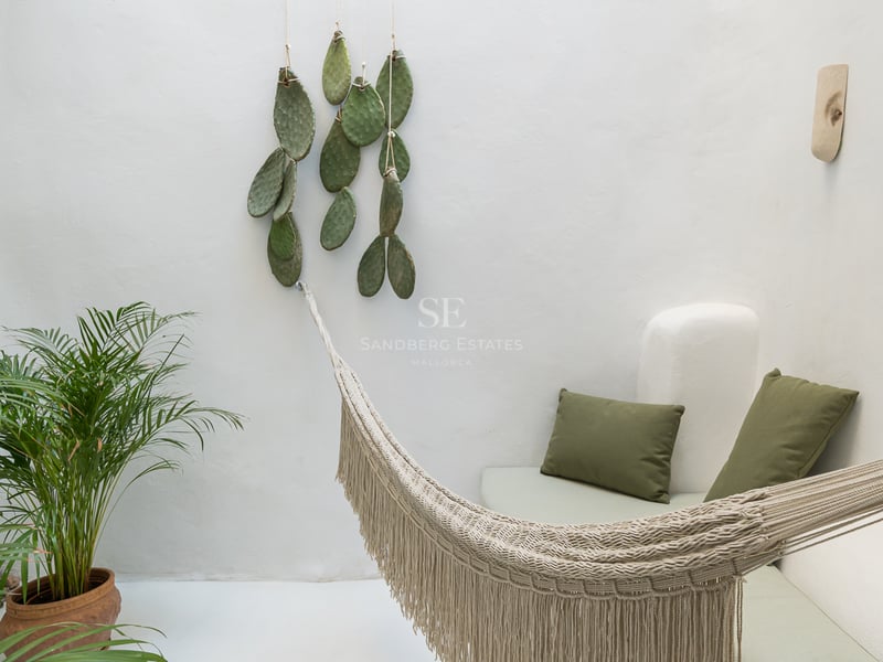 Un hamac tissé crème dans une cour minimaliste blanche avec des coussins verts et des plantes.