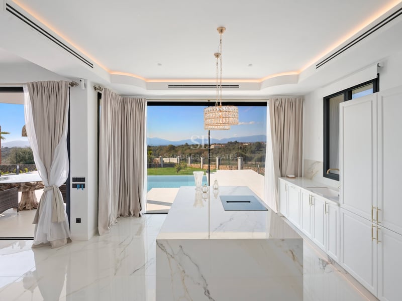 Cuisine lumineuse avec îlot central en marbre, placards blancs et baies vitrées ouvrant sur une piscine et la nature.
