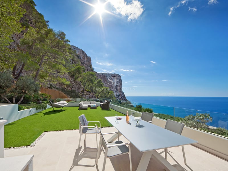 Terrasse moderne avec coin repas, pelouse artificielle et garde-corps en verre face à la mer et aux falaises.