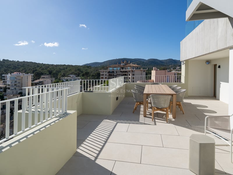 Terrasse moderne spacieuse avec table à manger en bois, chaises tressées et vue sur les collines environnantes.
