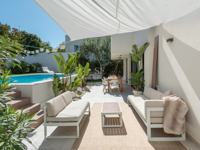 Terrasse moderne blanche avec canapés d'extérieur, coin repas et piscine turquoise sous une voile d'ombrage blanche.
