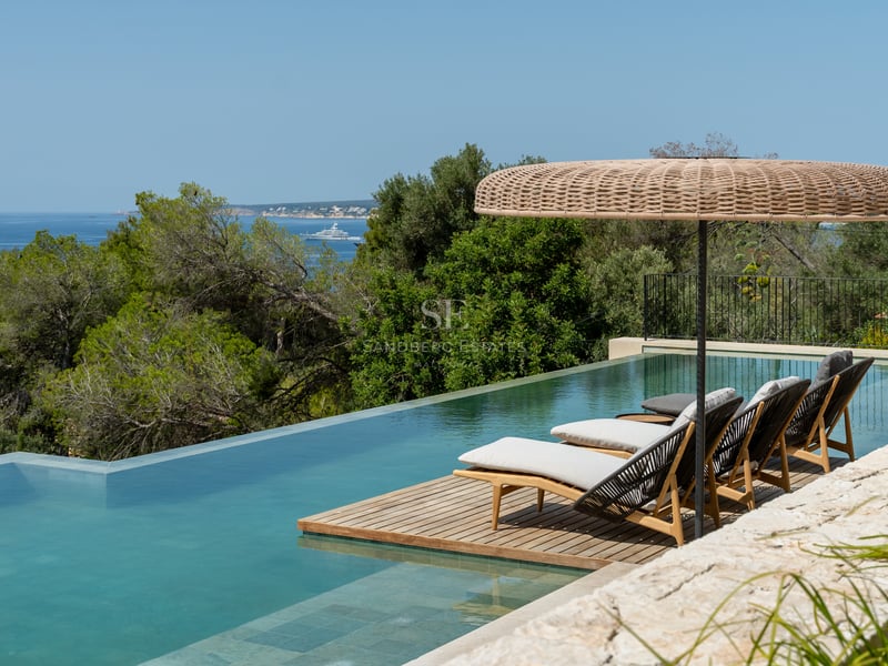 Piscine à débordement avec terrasse en bois, chaises longues et parasol en osier face à la mer et aux arbres.