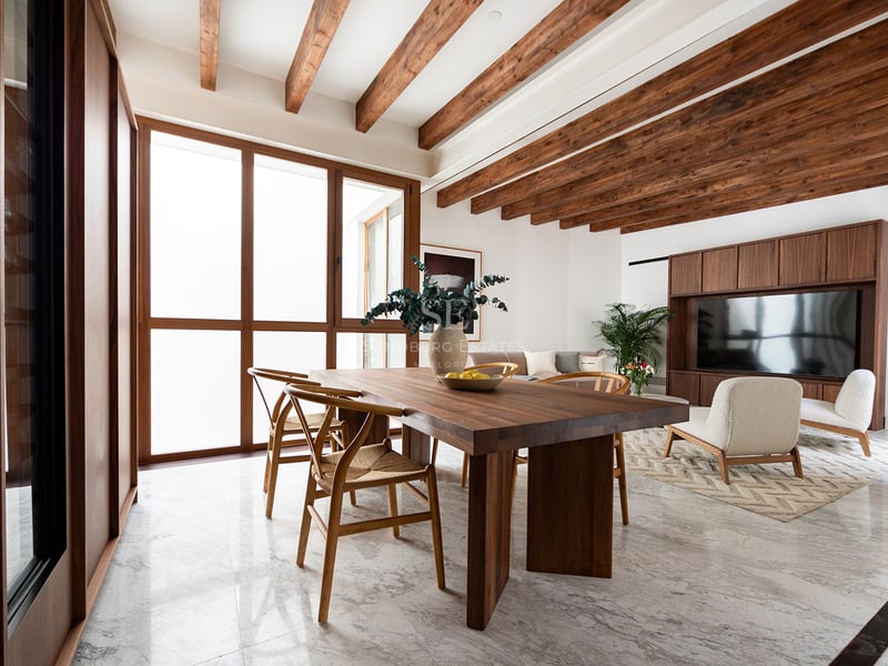 Salón moderno con vigas de madera, suelo de mármol, mesa de comedor de nogal y grandes ventanales.