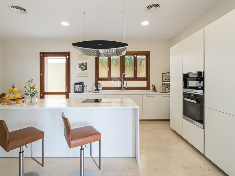 Cuisine blanche moderne avec îlot central, tabourets en cuir, sol en marbre et cadres de fenêtres en bois.