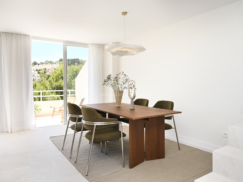 Salle à manger moderne avec table en bois, chaises vert olive et grandes baies vitrées ouvrant sur une terrasse ensoleillée.