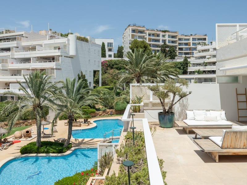Terrasse ensoleillée avec mobilier en bois surplombant des piscines bleues et des bâtiments blancs méditerranéens.