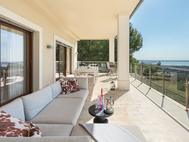 Terrasse de luxe couverte avec sol en pierre naturelle, canapé gris, garde-corps en verre et vue sur la mer et la forêt.