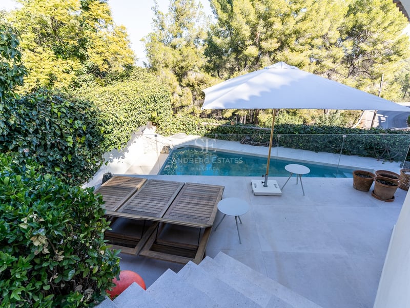 Terrasse en pierre avec piscine privée, chaises longues en bois et parasol blanc entourée de pins.