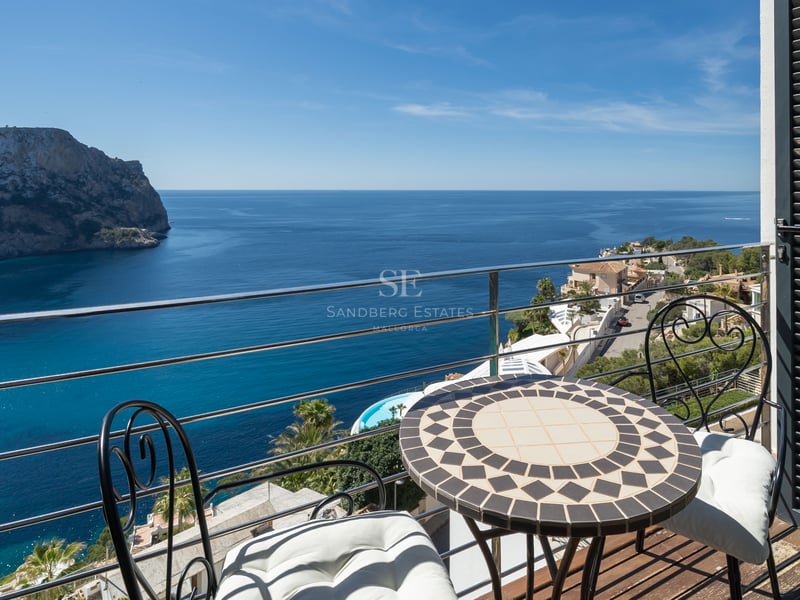 Une petite terrasse avec table en mosaïque et chaises en fer donnant sur une baie méditerranéenne bleue et des falaises.