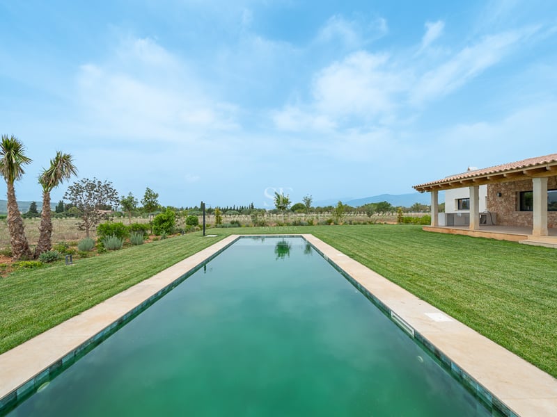 Longue piscine rectangulaire centrée sur une vaste pelouse verte avec une villa en pierre et des montagnes au loin.