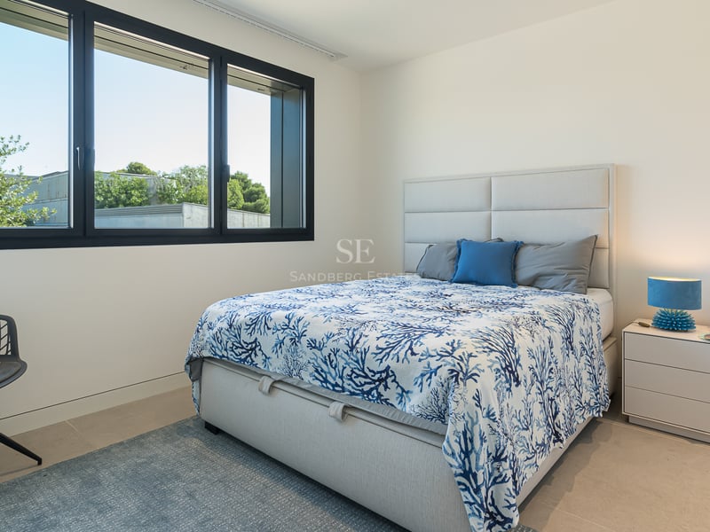 Chambre contemporaine avec grande fenêtre, lit aux motifs bleus, table de chevet blanche et chaise design.